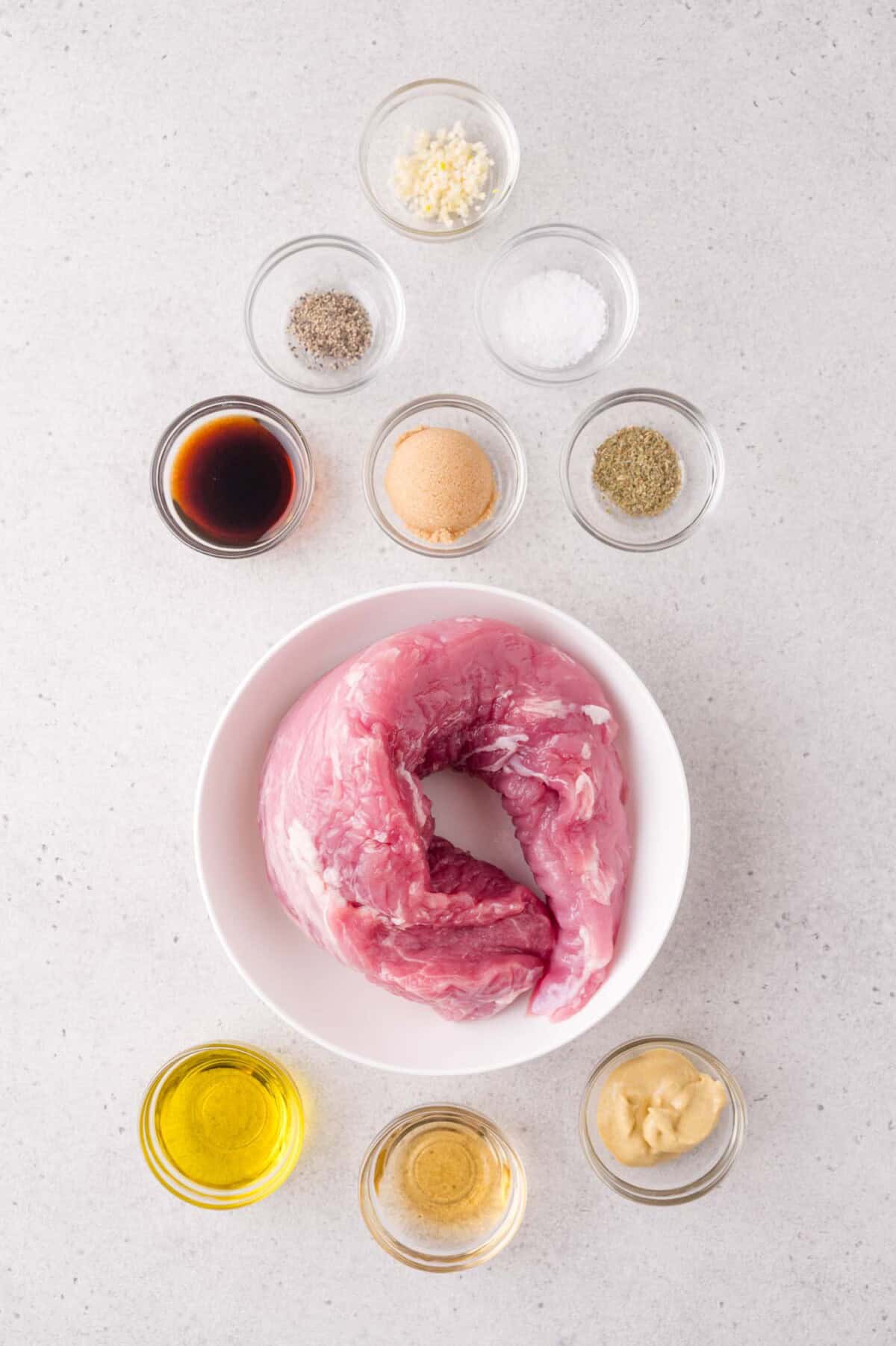 Raw pork tenderloin in a white bowl surrounded by small bowls of seasoning, oil, soy sauce, mustard, brown sugar, and minced garlic on a light surface.
