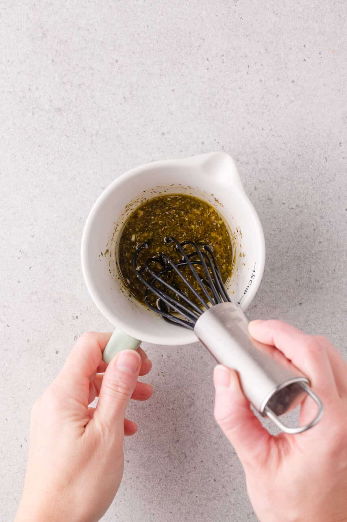 A person whisking marinade in a white ceramic measuring cup on a light gray surface.