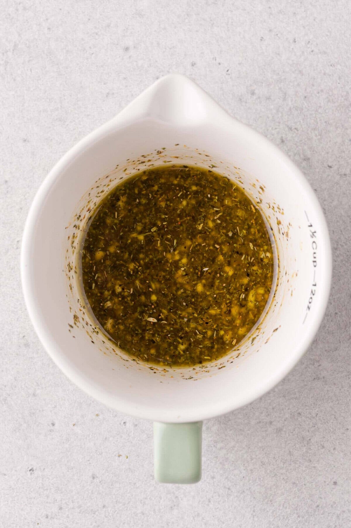 A white measuring cup filled with a mixed herb and oil marinade sits on a light gray surface.