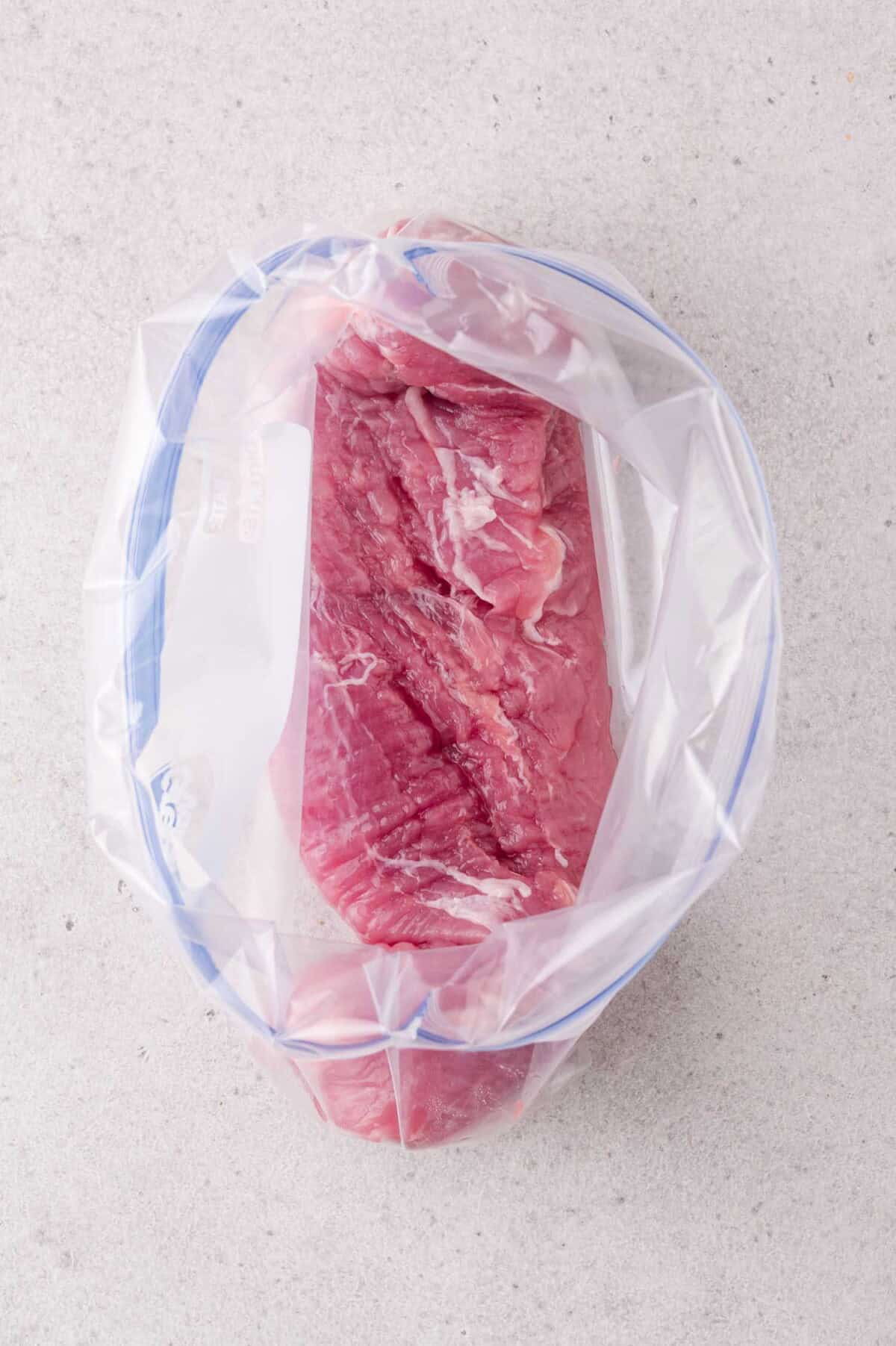 Raw pork tenderloin placed inside a clear resealable plastic bag, set on a light-colored surface.