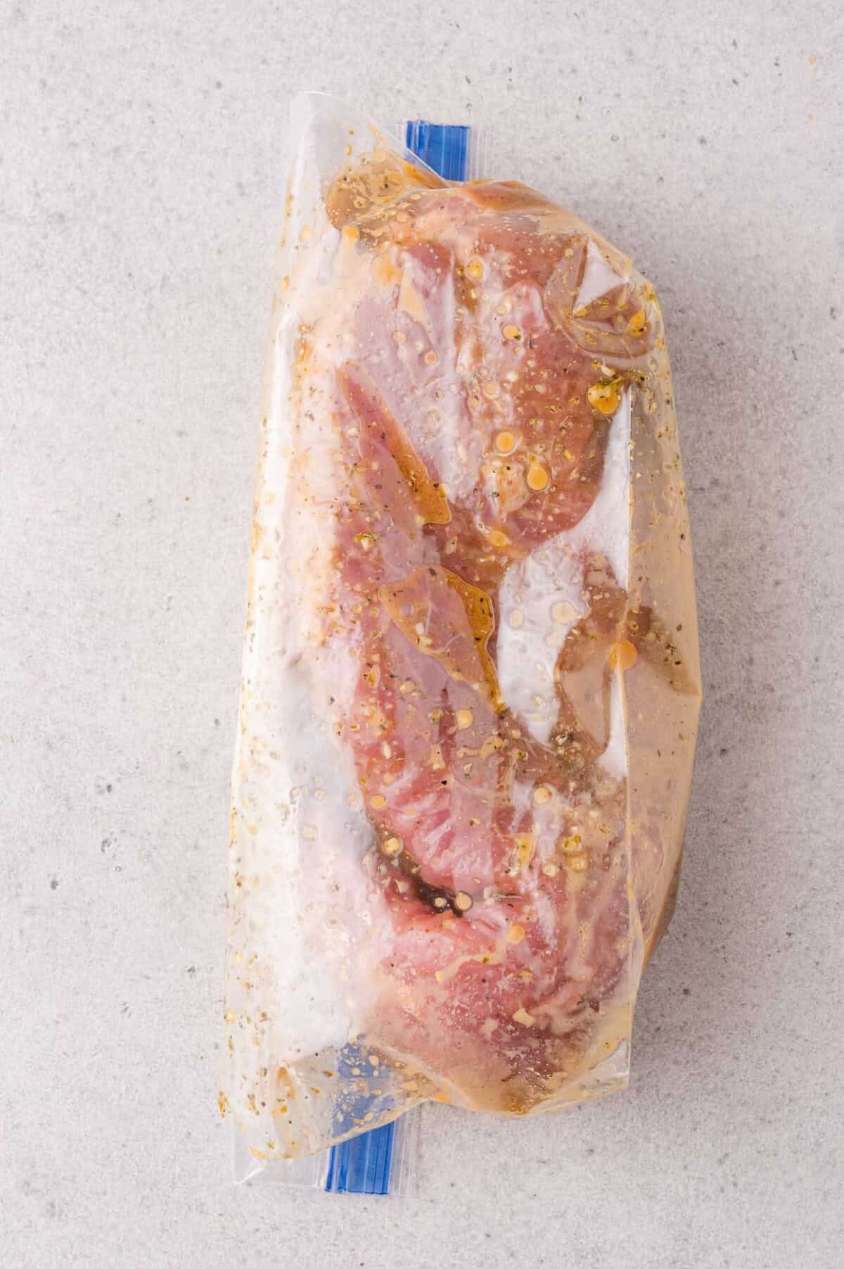 Raw marinated meat sealed in a clear plastic ziplock bag, placed on a light gray textured surface.