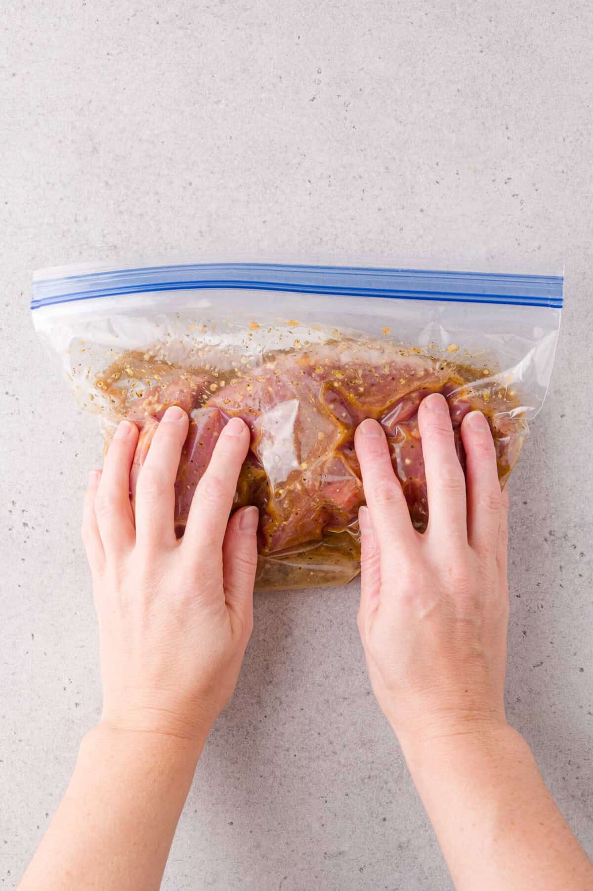 Hands sealing a plastic zip-top bag containing marinated meat on a light-colored countertop.