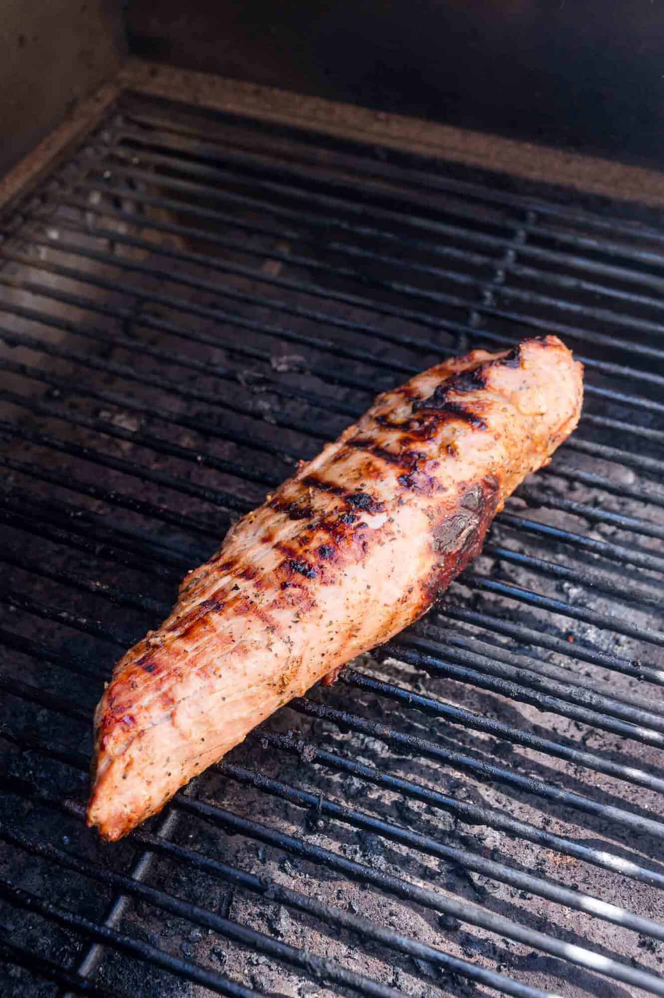 A seasoned pork tenderloin cooking on a grill, showing visible grill marks and slight charring.