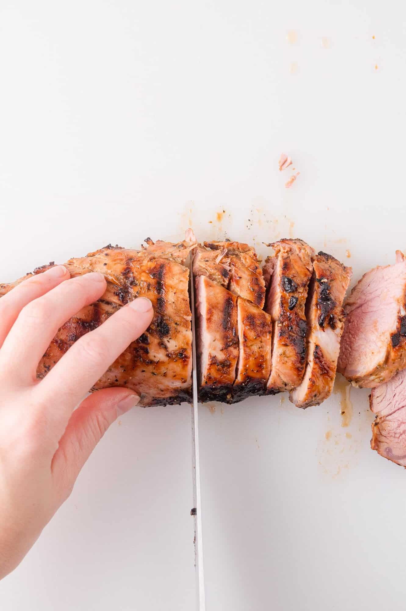 A hand slices cooked pork tenderloin into pieces on a white cutting board.