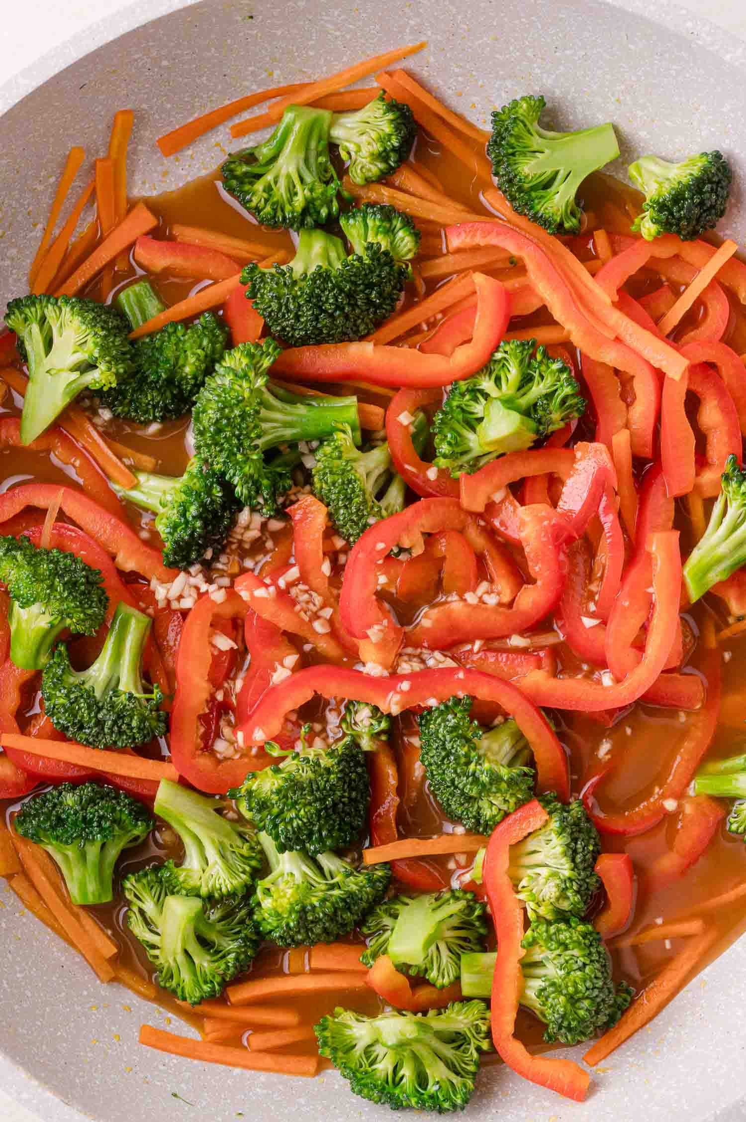 Broccoli florets, sliced red bell peppers, and carrots cooking in a pan with a brown sauce and minced garlic.