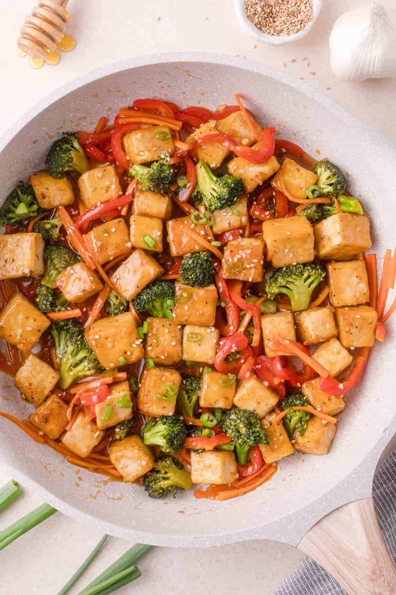 A stir-fry dish with cubed tofu, broccoli, red bell peppers, and carrots in a light sauce, topped with sesame seeds, served in a pan.