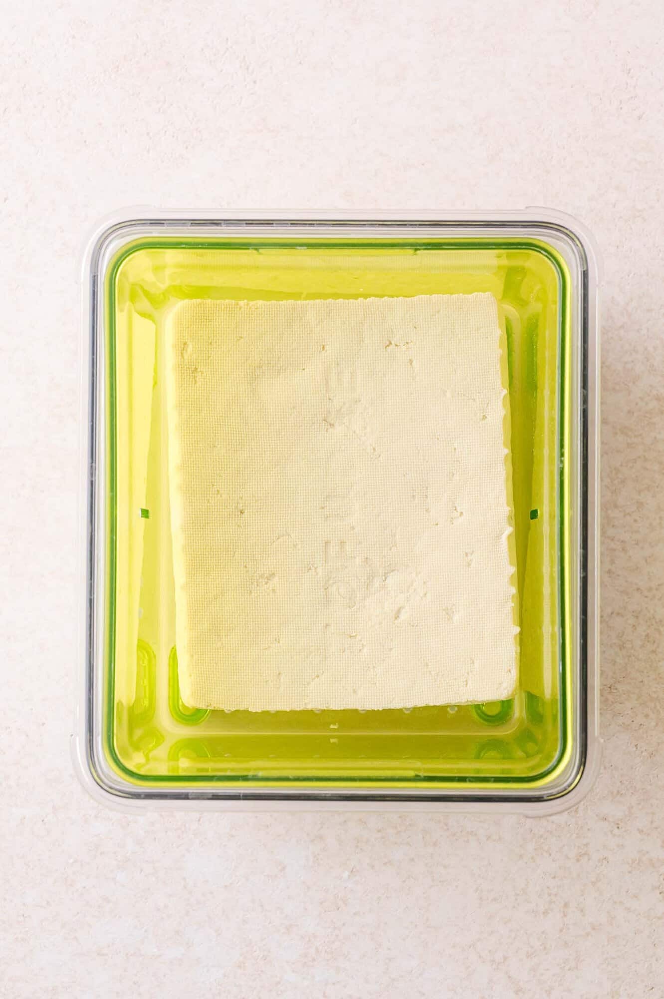 A block of tofu sits inside a green plastic container, viewed from above on a light-colored surface.