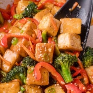 Stir-fried tofu cubes with broccoli and red bell pepper, garnished with sesame seeds and sliced green onions, served with a black spatula.