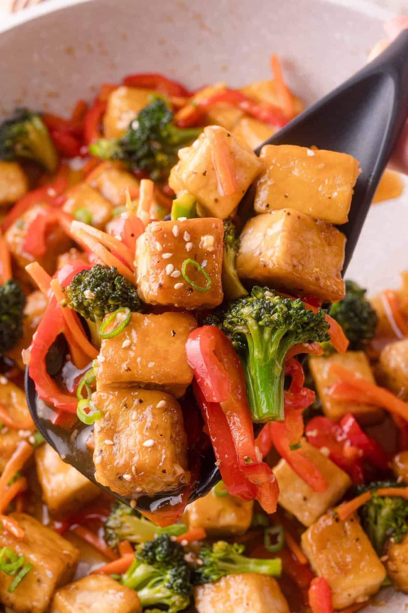 A close-up of a serving spoon holding tofu cubes, broccoli, red bell pepper, and carrot strips in a savory sauce.