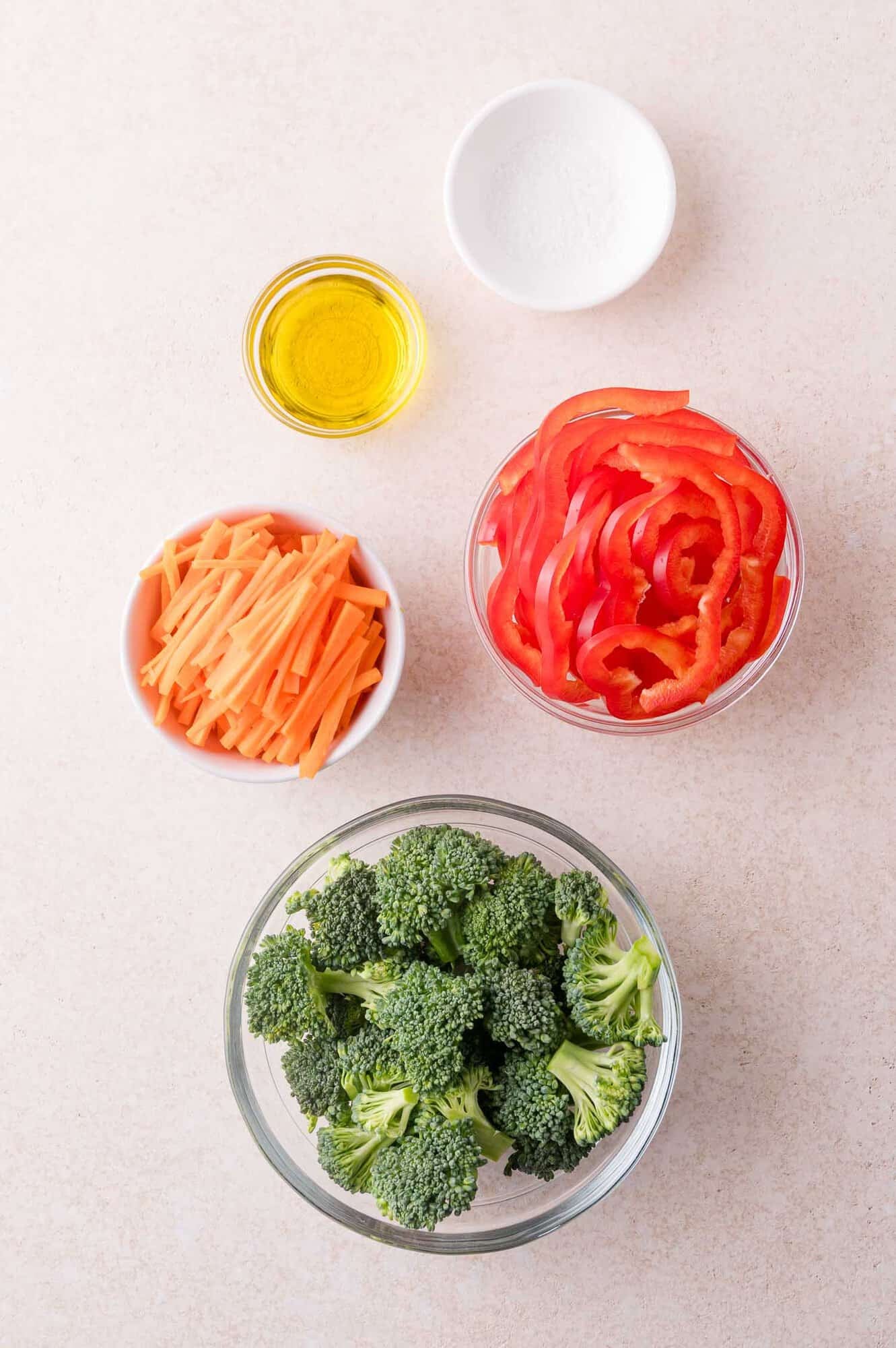 Glass bowls with broccoli florets, sliced red bell pepper, julienned carrots, a small bowl of olive oil, and a small bowl of salt on a light surface.