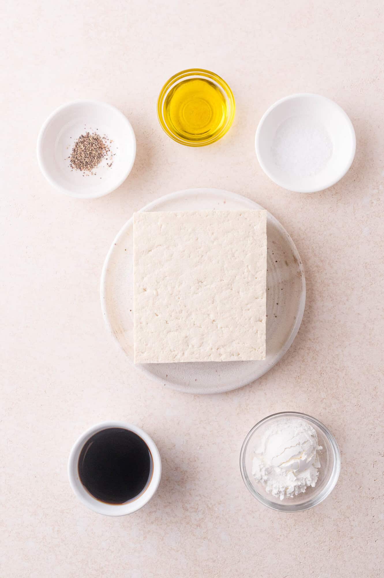 A block of tofu on a plate surrounded by small bowls containing black pepper, olive oil, salt, soy sauce, and cornstarch on a light surface.