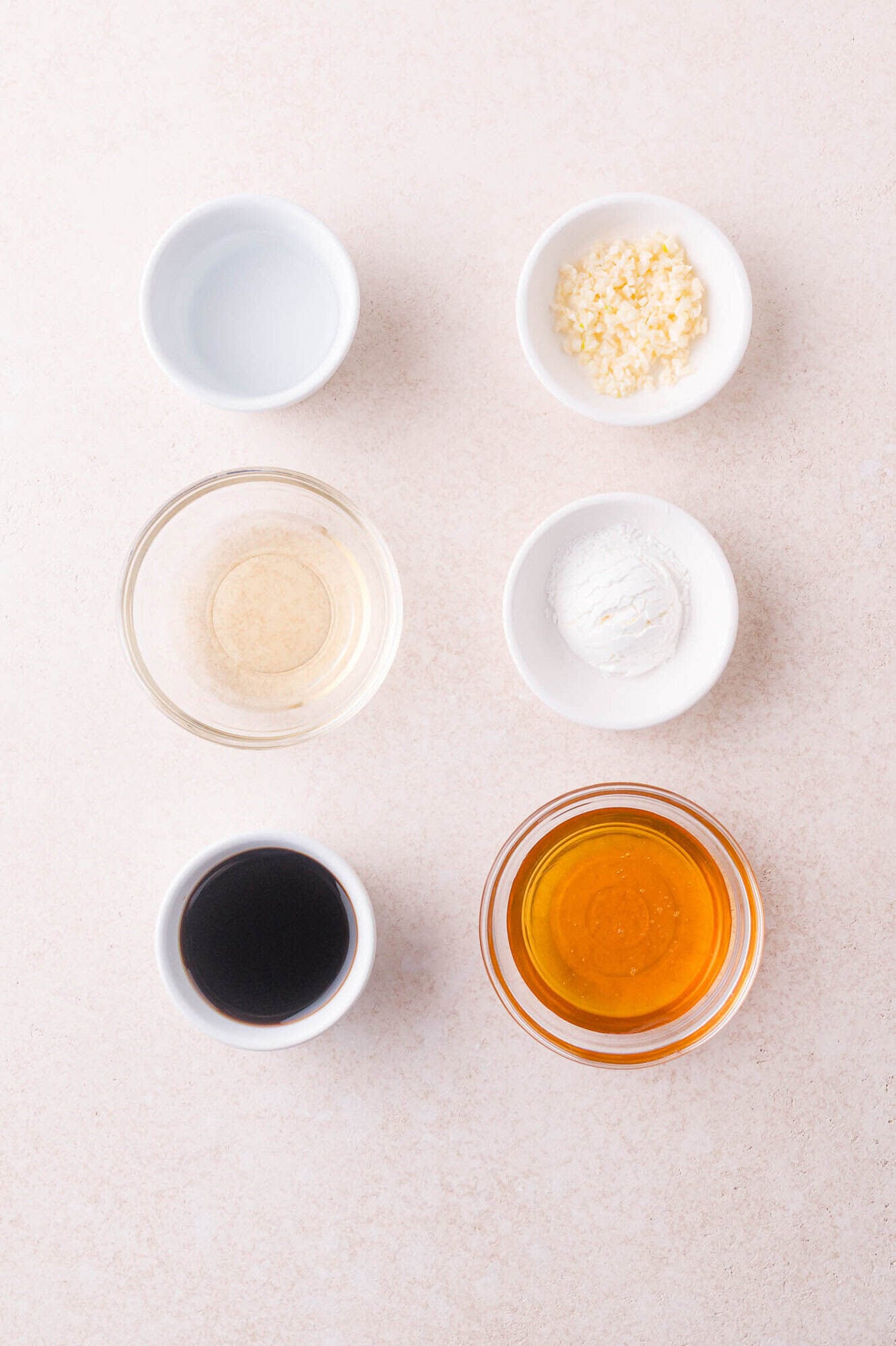 Six small bowls with various ingredients: chopped garlic, cornstarch, honey, soy sauce, rice vinegar, and water, arranged on a light-colored surface.