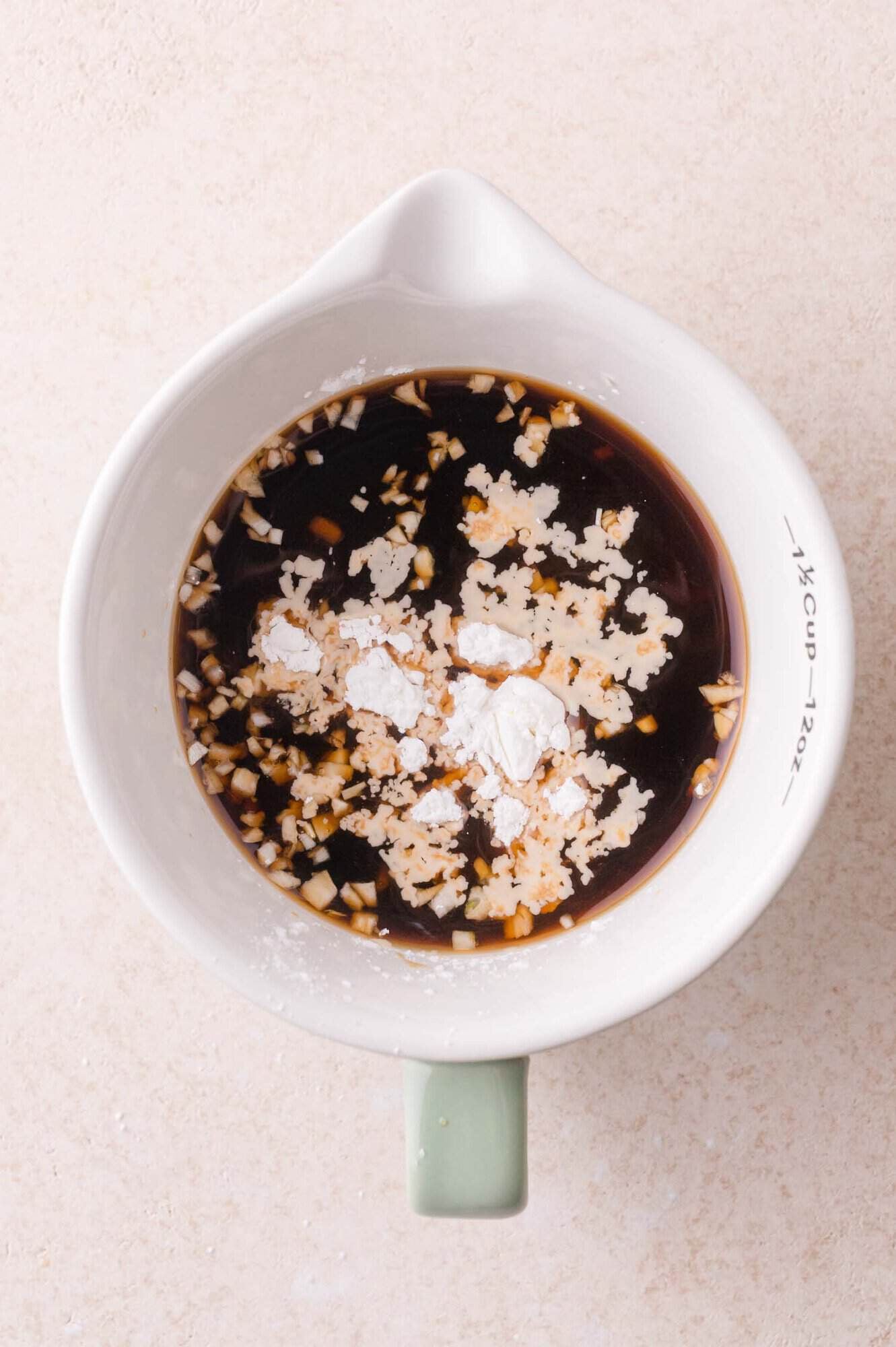 A white measuring cup with soy sauce, minced garlic, and cornstarch mixed together on a light countertop.