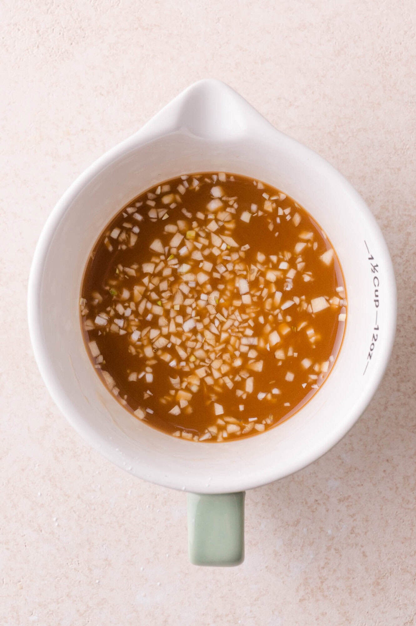A white measuring cup filled with a brown liquid containing finely chopped garlic, placed on a light beige surface.
