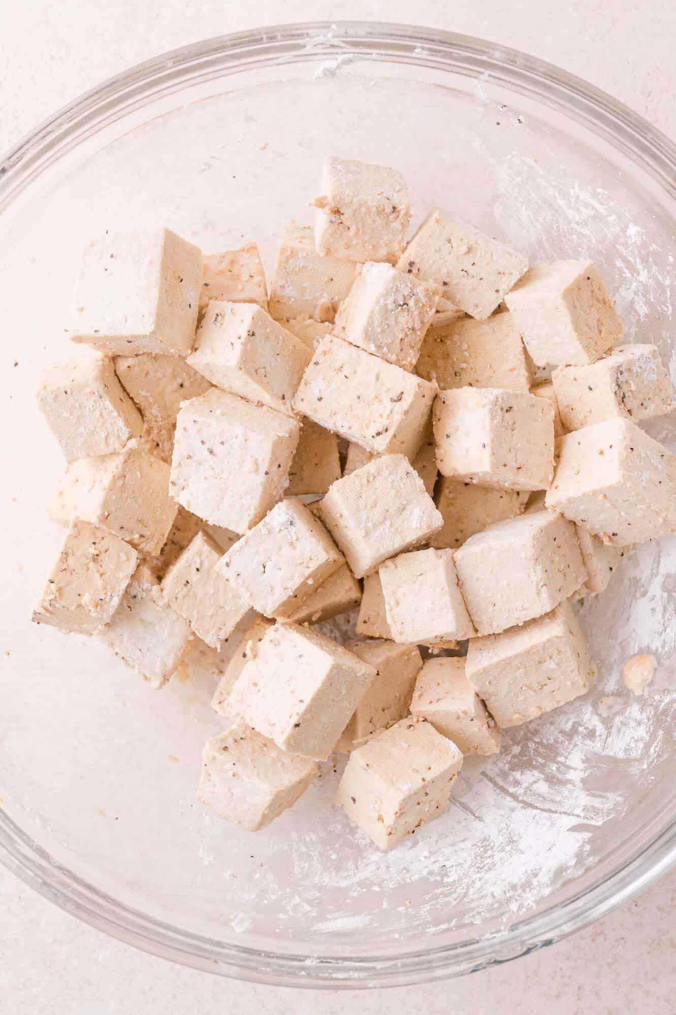 Cubed tofu coated with seasoning in a clear glass mixing bowl, set on a light-colored surface.