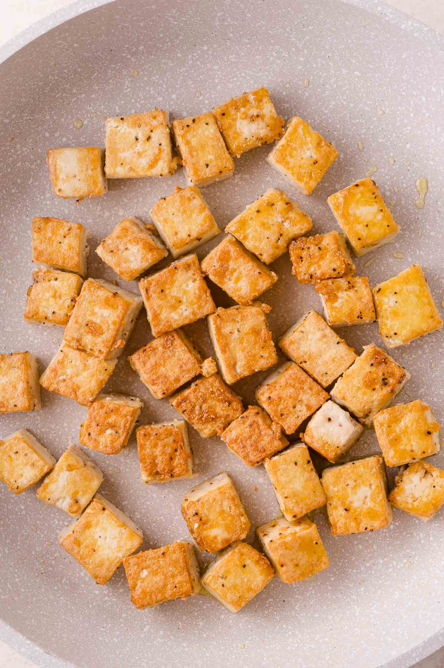 Cubed tofu is pan-fried to a golden brown in a light-colored skillet.