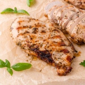 Grilled chicken breast with Italian herb seasoning on parchment paper, with fresh basil leaves and sliced chicken in the background.