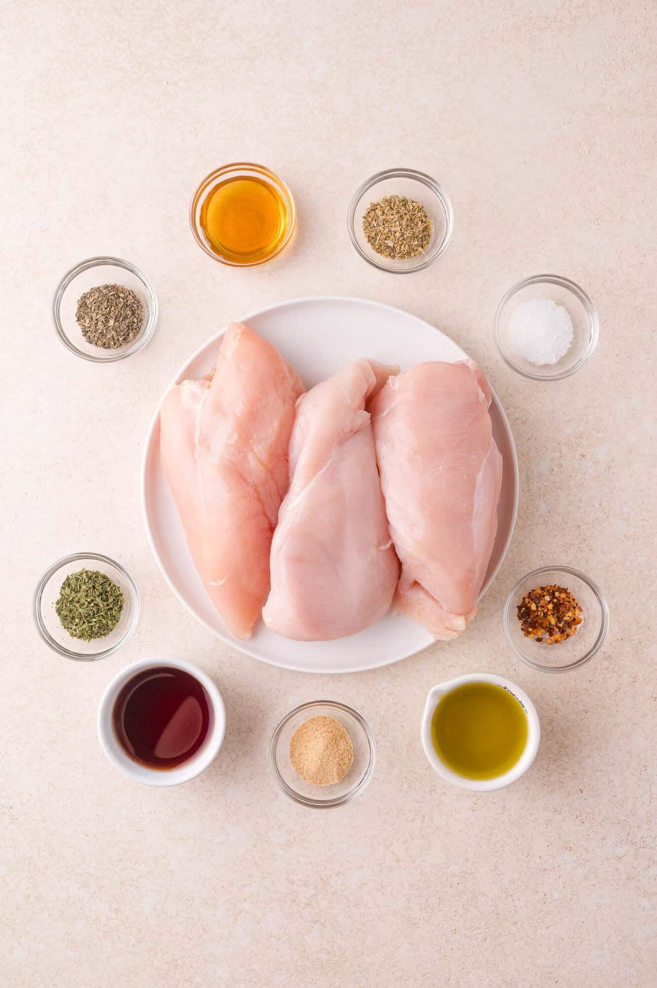 Three raw chicken breasts on a white plate, surrounded by small bowls of various spices, seasonings, and liquids on a light surface.