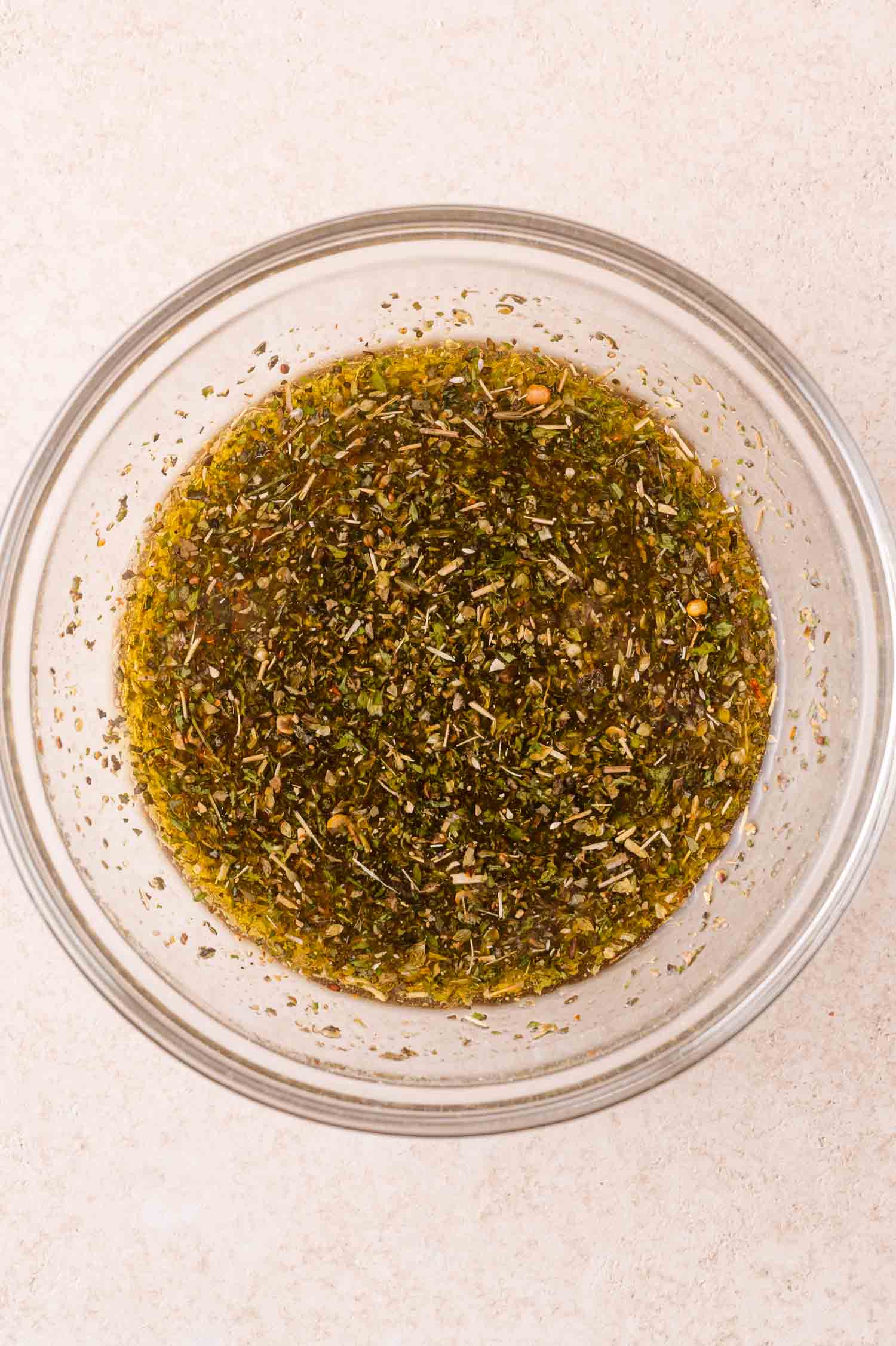 A clear glass bowl filled with a mixture of olive oil and various dried herbs and spices on a light-colored surface.