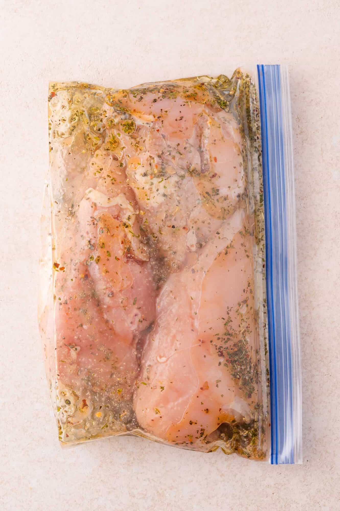 Two raw chicken breasts marinating in herbs and seasoning inside a sealed plastic zipper bag on a light surface.