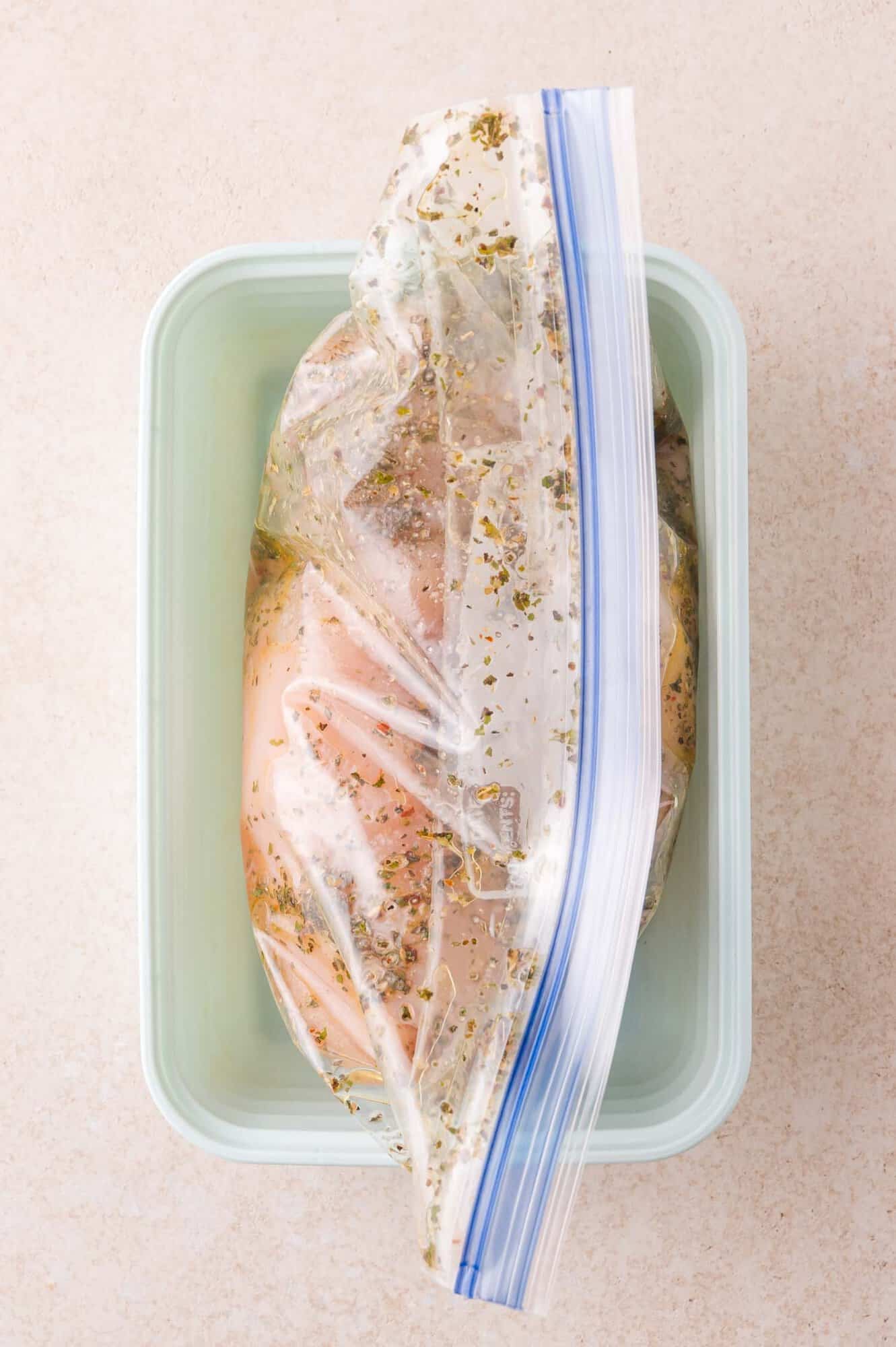 Raw chicken breast marinating in Italian marinade inside a sealed plastic bag, placed in a rectangular container on a light-colored surface.