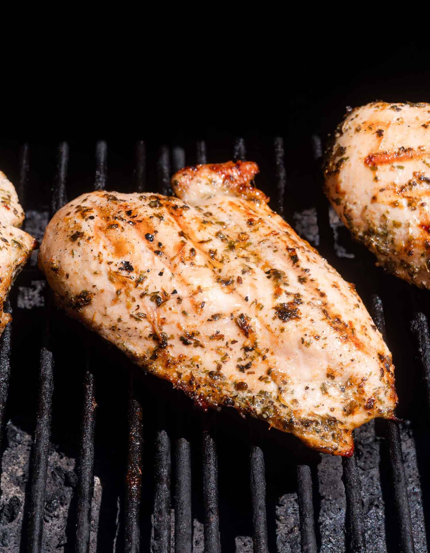Seasoned chicken breast grilling on a barbecue, with visible grill marks and herbs on the surface.