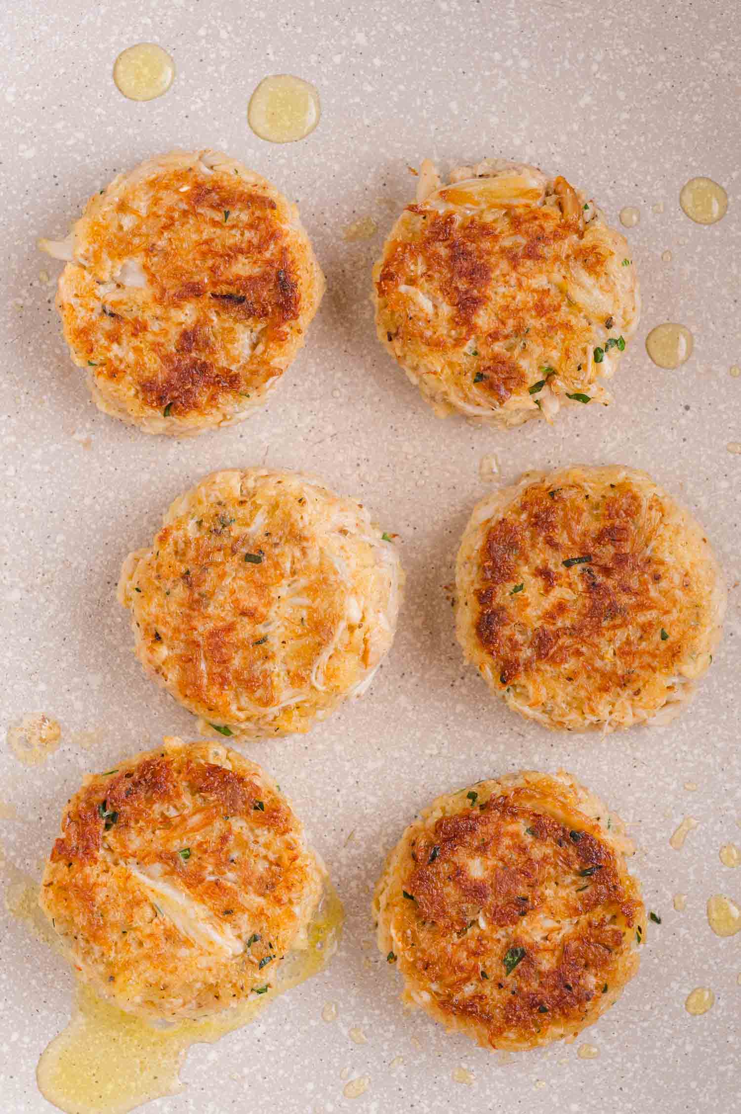 Six golden-brown crab cakes are arranged in a skillet with small drops of oil around them.