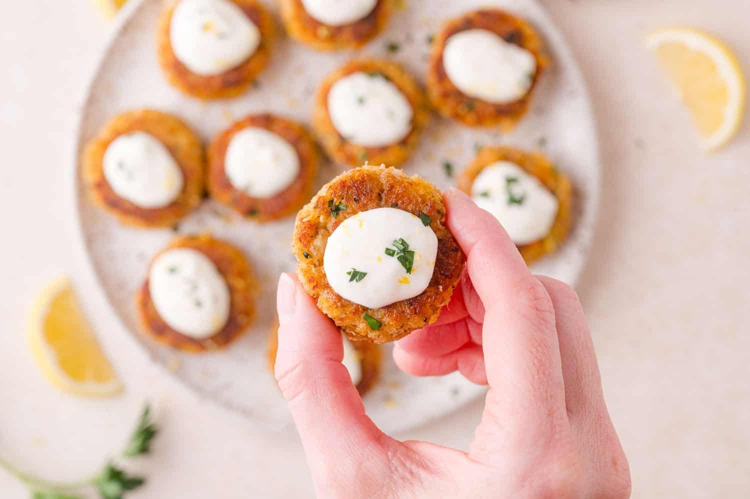 A hand holding a small, round crab cake topped with aioli and herbs, with more appetizers on a plate in the background. Lemon wedges and herbs are scattered nearby.