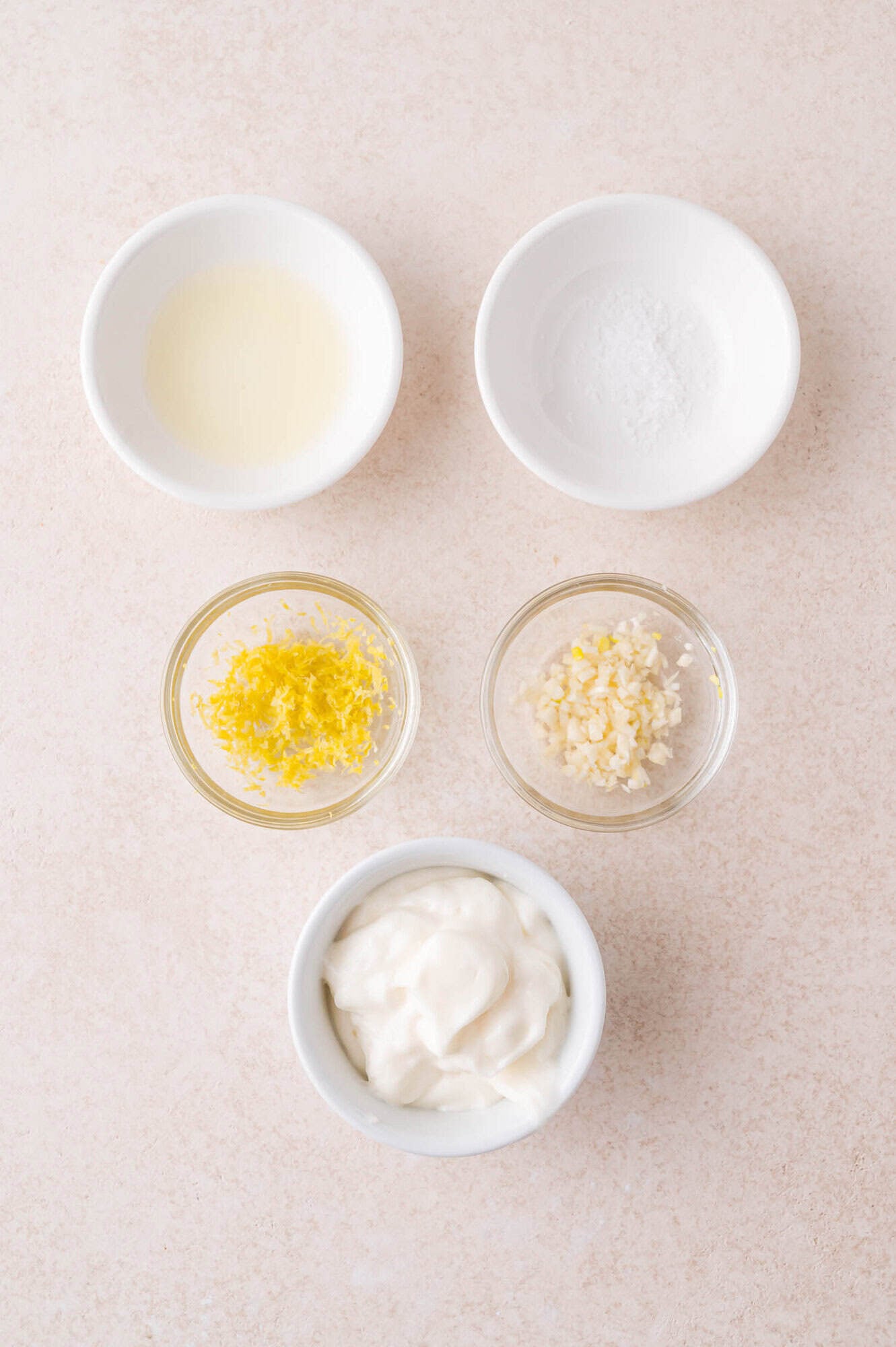 Five small bowls containing lemon juice, salt, lemon zest, minced garlic, and mayonnaise arranged on a light surface.
