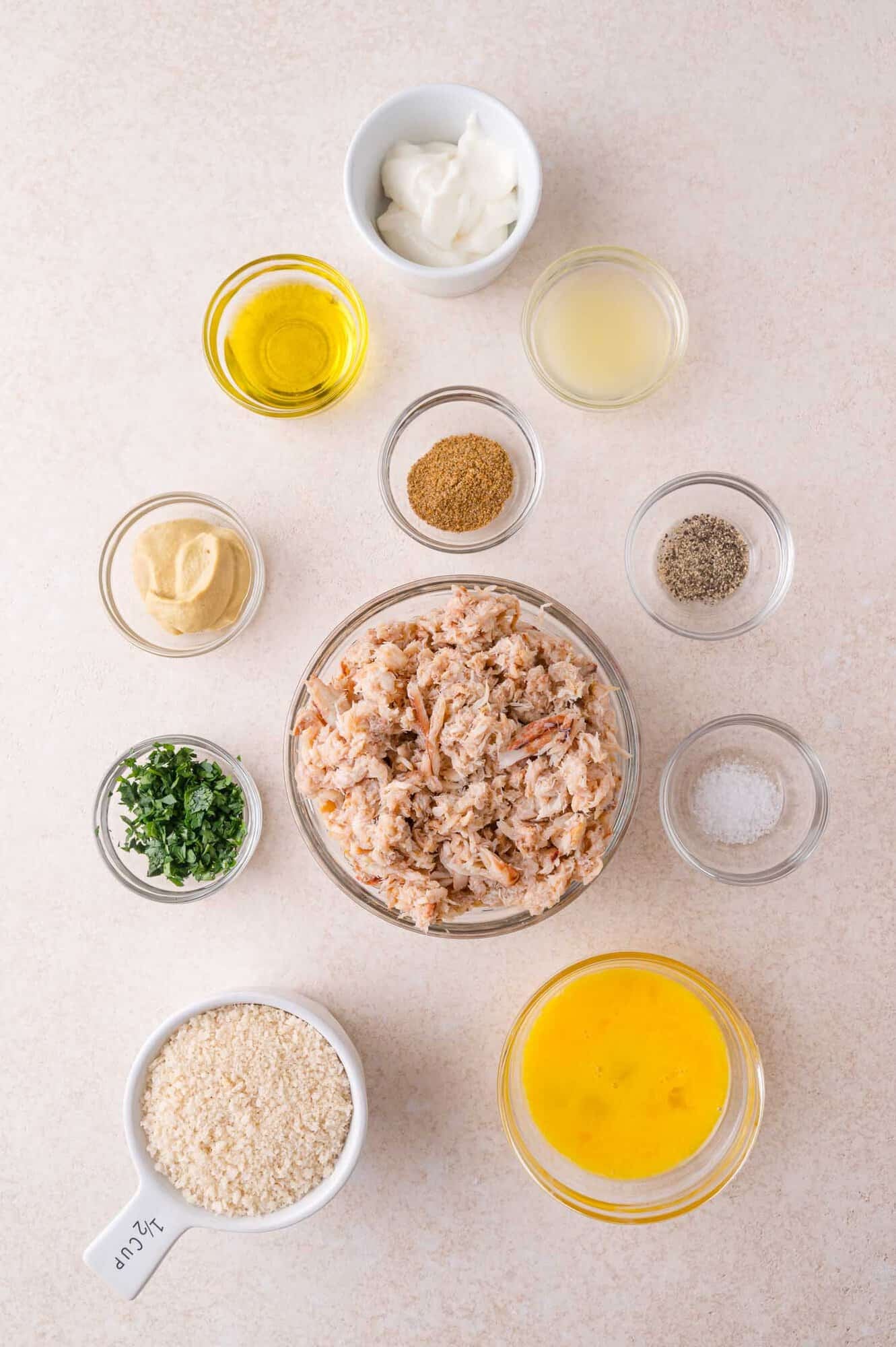 Overhead view of separate bowls containing crab meat, panko breadcrumbs, egg, mayonnaise, mustard, parsley, oil, lemon juice, and seasonings arranged on a light surface.