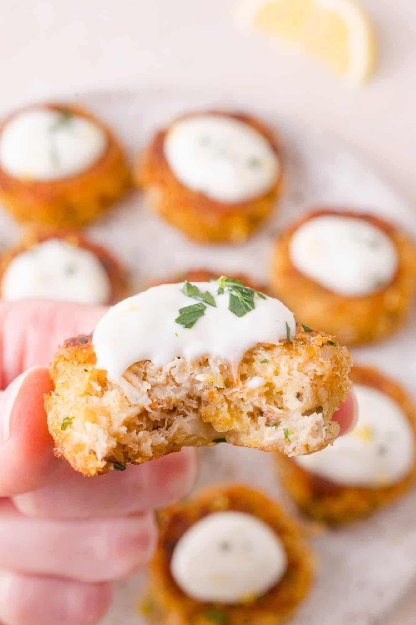 A hand holds a partially eaten mini crab cake topped with white aioli; several whole crab cakes with sauce are visible in the background.