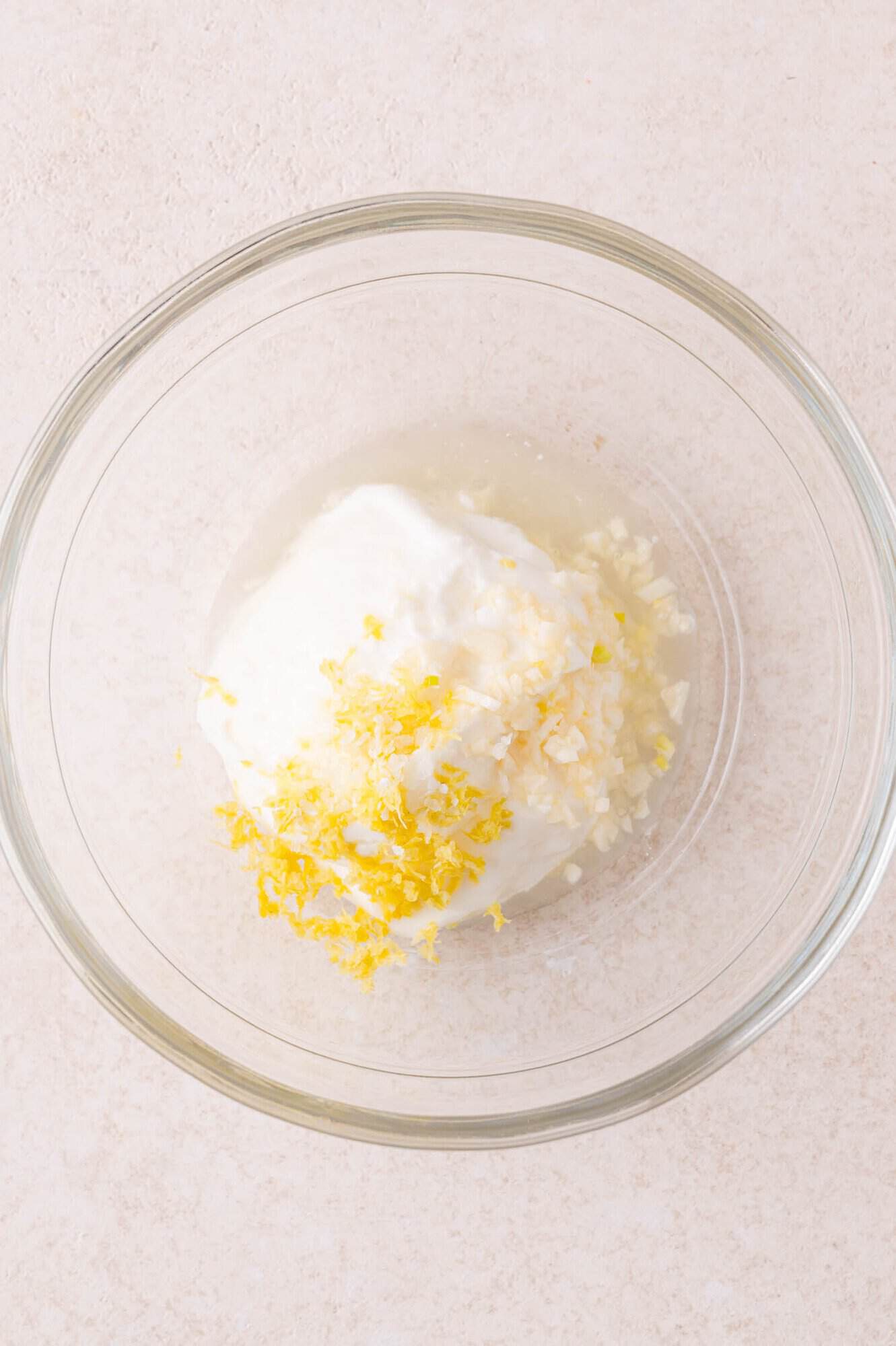 A glass bowl containing yogurt, minced garlic, lemon zest, and lemon juice on a light-colored surface.