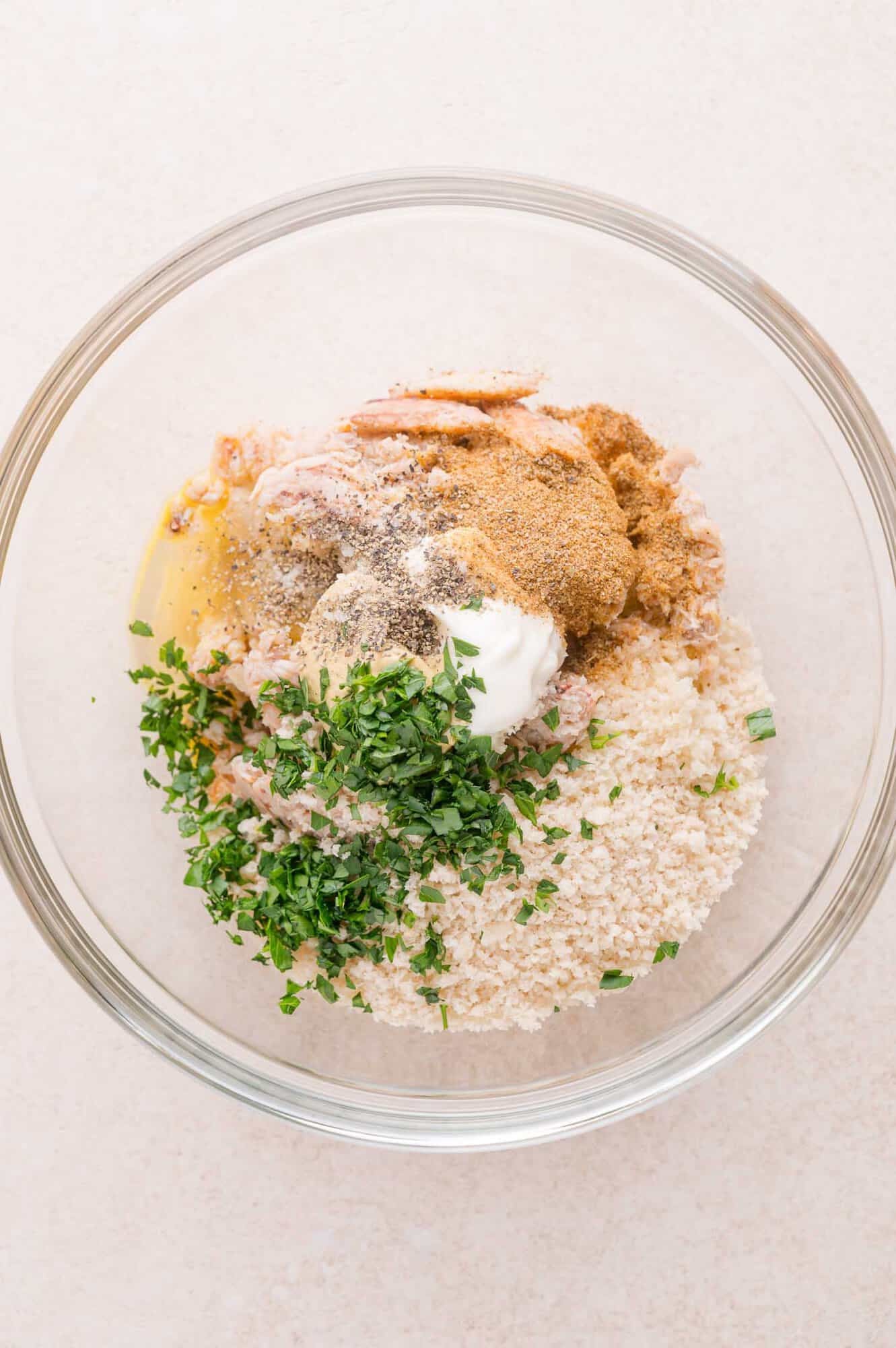 A glass bowl containing crab meat, breadcrumbs, chopped herbs, seasoning, an egg, and a dollop of mayonnaise on a light surface.