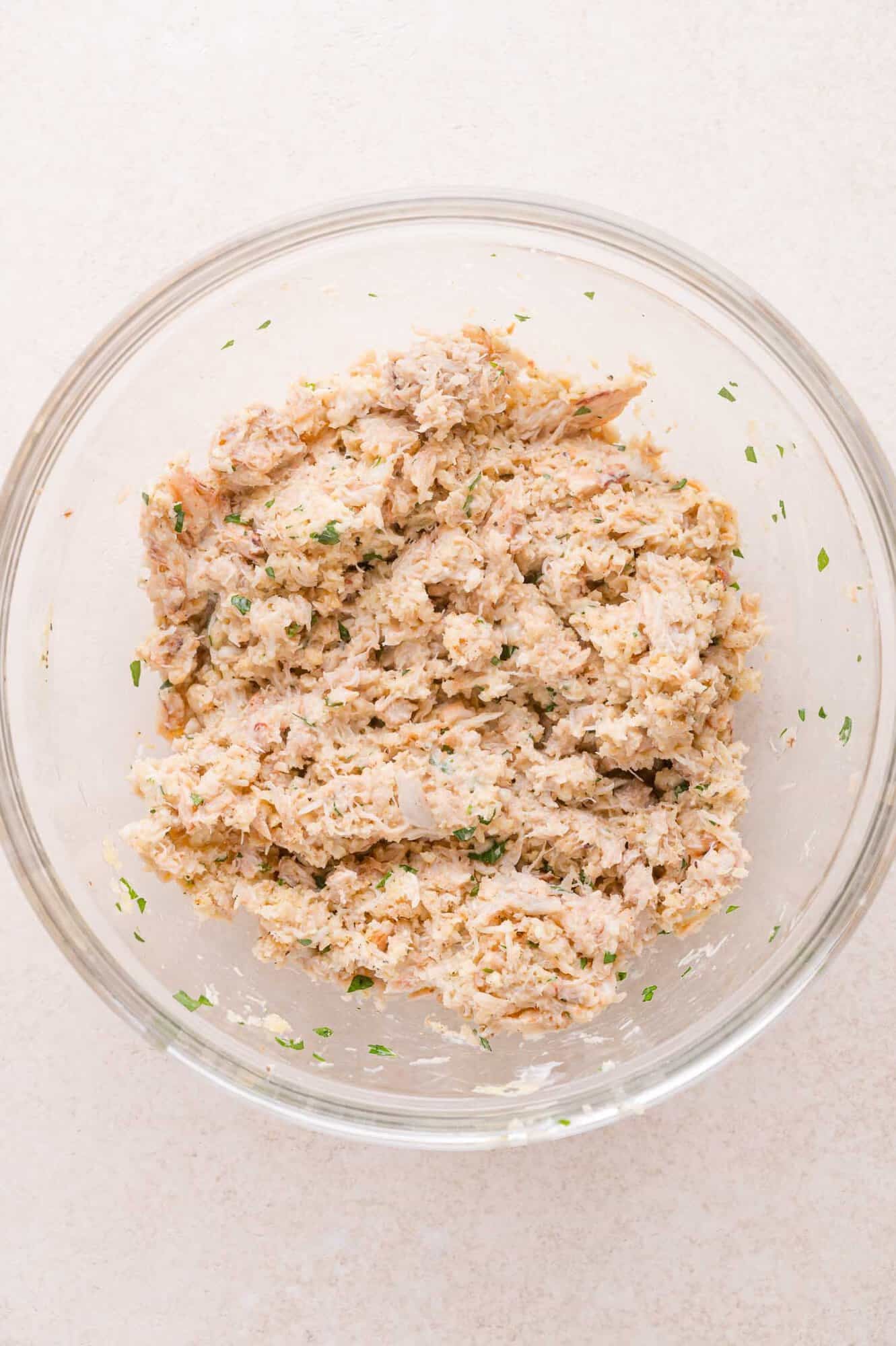 A clear glass bowl containing a mixture of crab, herbs, and other finely chopped ingredients on a light surface.