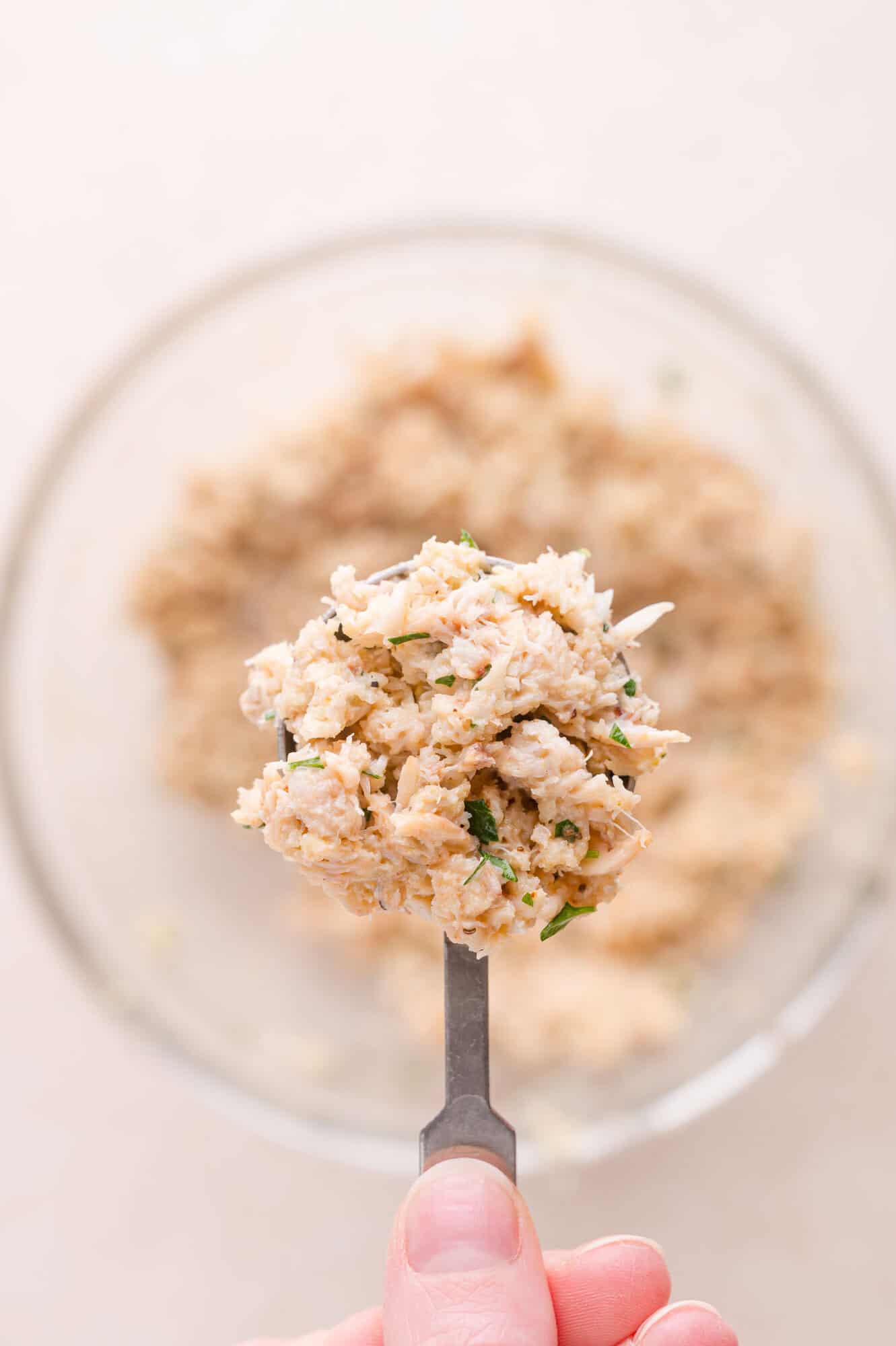 A hand holds a spoonful of a mixed crab filling above a glass bowl containing more of the mixture.