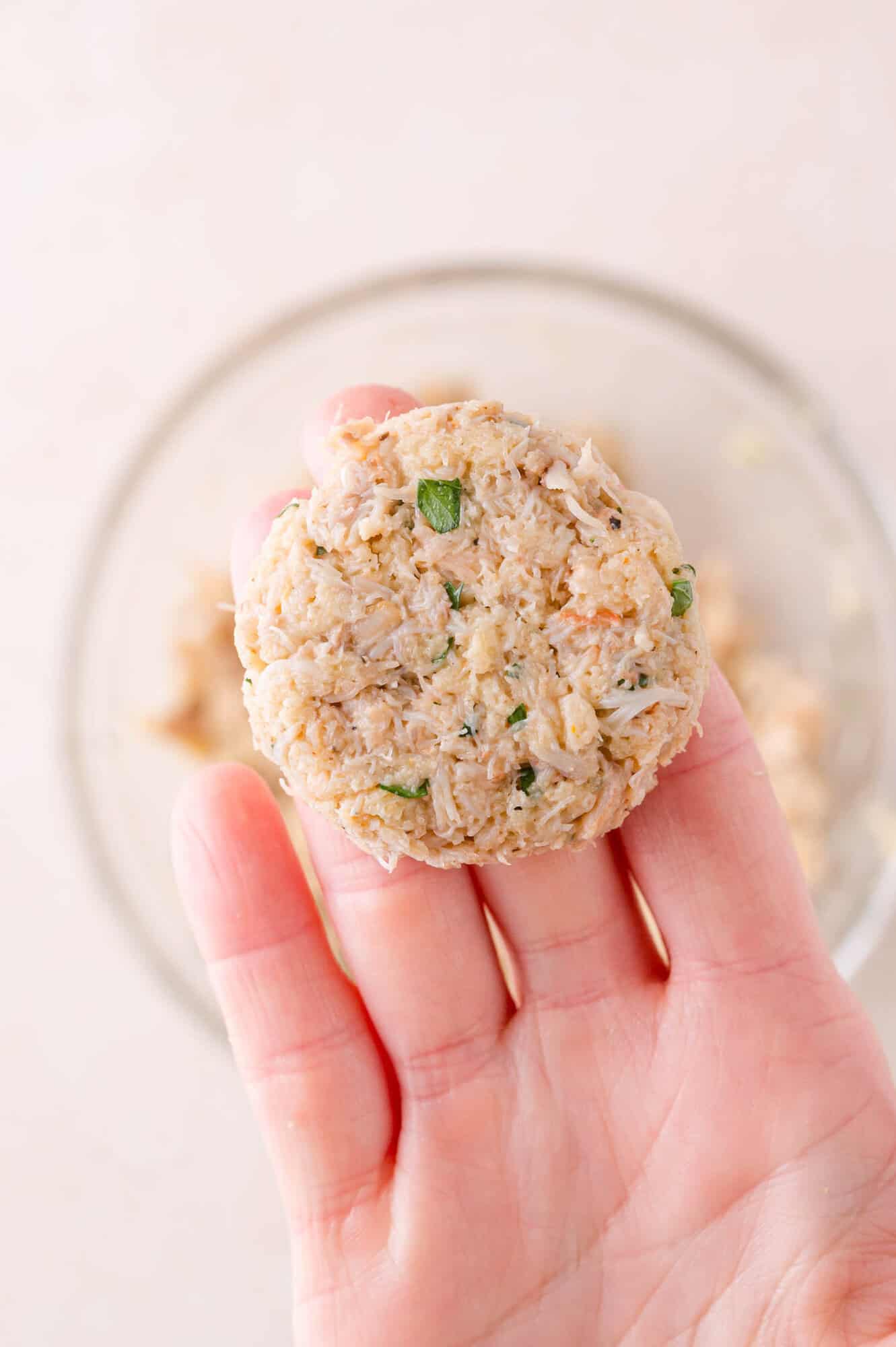 A hand holds an uncooked crab cake patty above a glass bowl containing the crab mixture.
