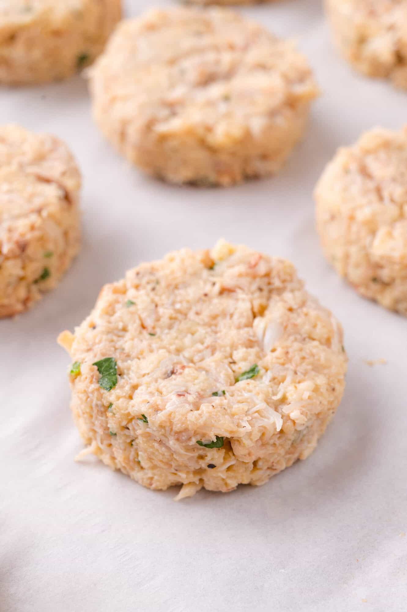 Uncooked crab cakes formed into round patties rest on a sheet of parchment paper.