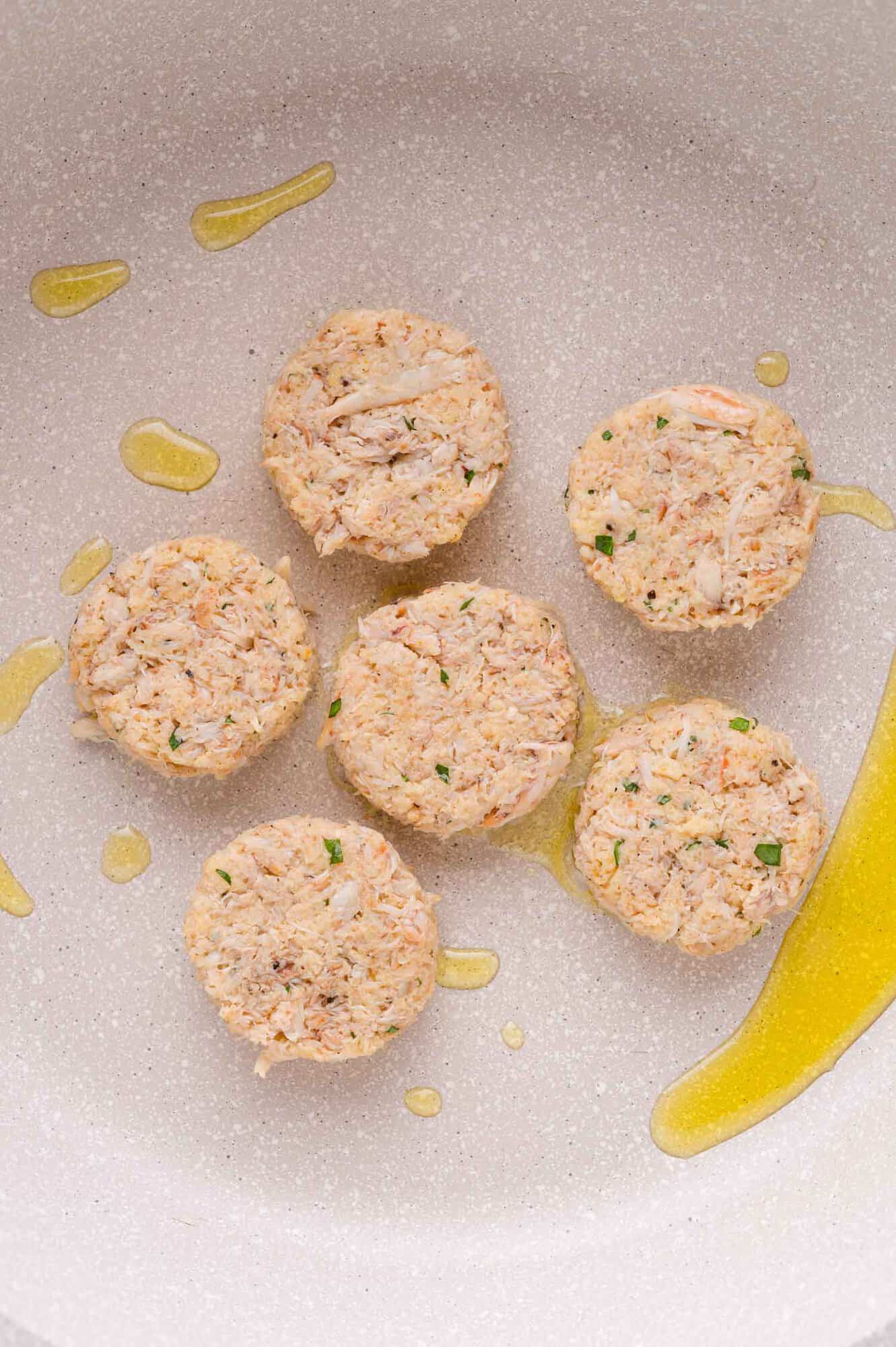 Six uncooked crab cakes arranged in a frying pan with a drizzle of oil.