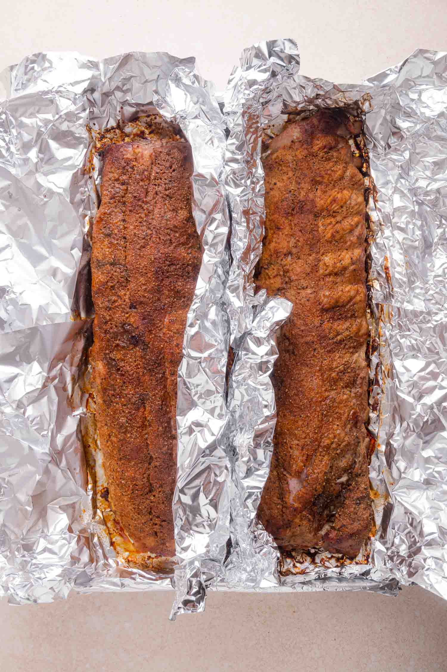Two racks of seasoned, cooked ribs resting in aluminum foil on a light-colored surface.