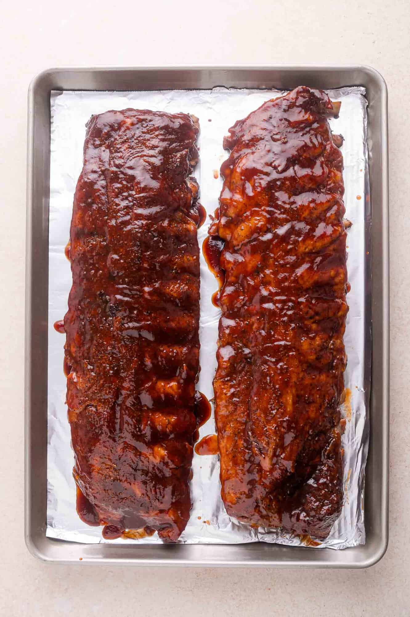 Two racks of barbecued ribs covered in sauce on a foil-lined baking tray.