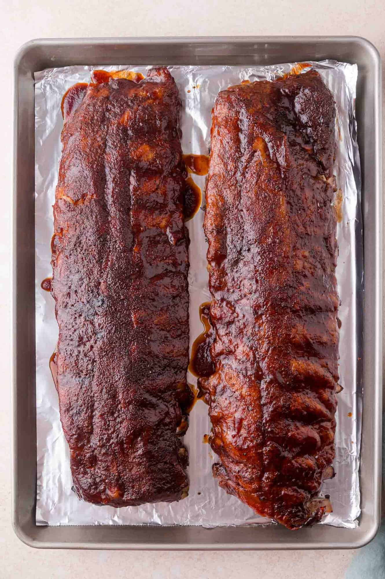 Two racks of cooked, glazed ribs rest on a foil-lined baking sheet.