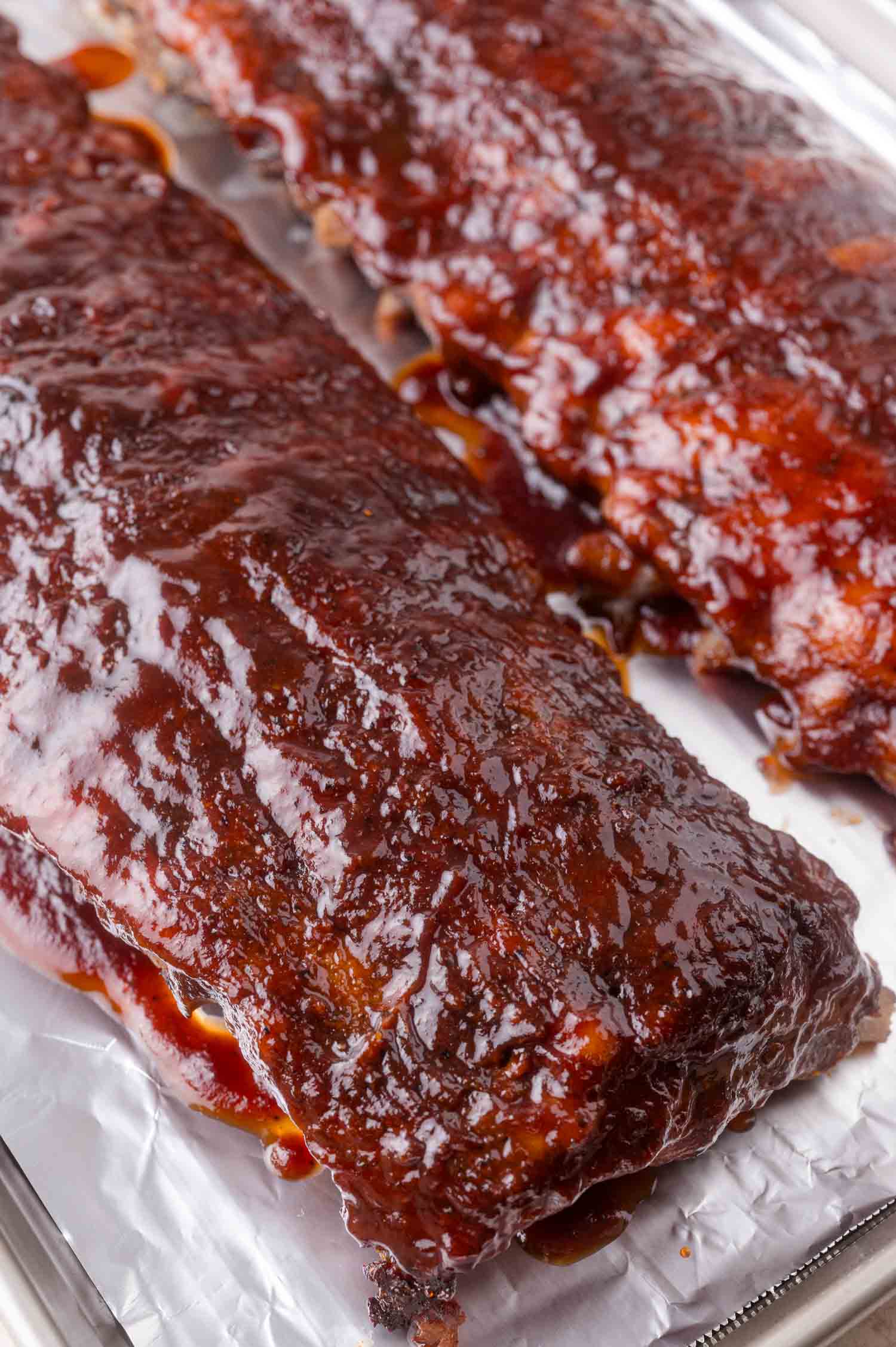 Two racks of barbecued ribs covered in glossy sauce rest on a foil-lined baking sheet.