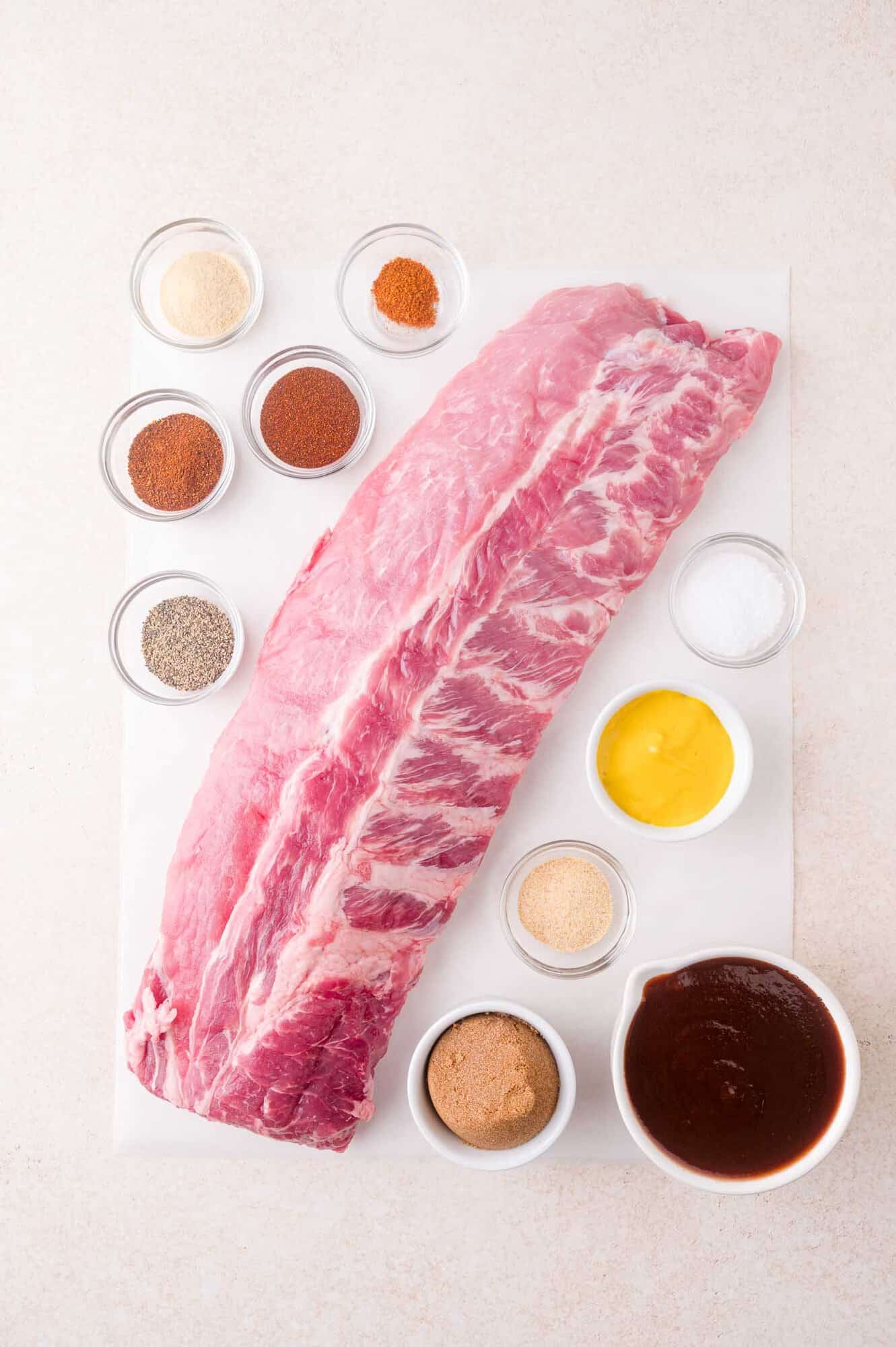 Uncooked rack of pork ribs on a white surface, surrounded by small bowls of spices, mustard, salt, pepper, sugar, and barbecue sauce.