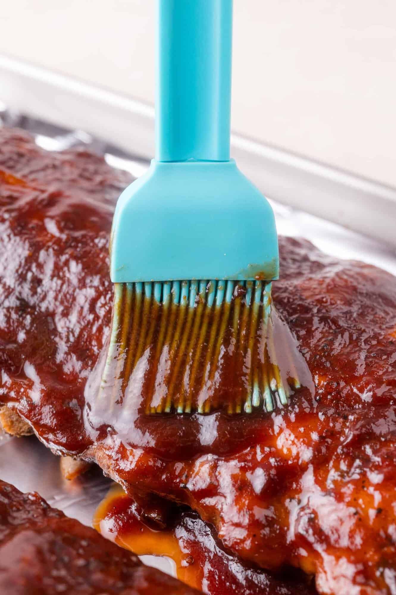 A close-up of a blue silicone brush applying barbecue sauce onto a rack of ribs on a metal tray.