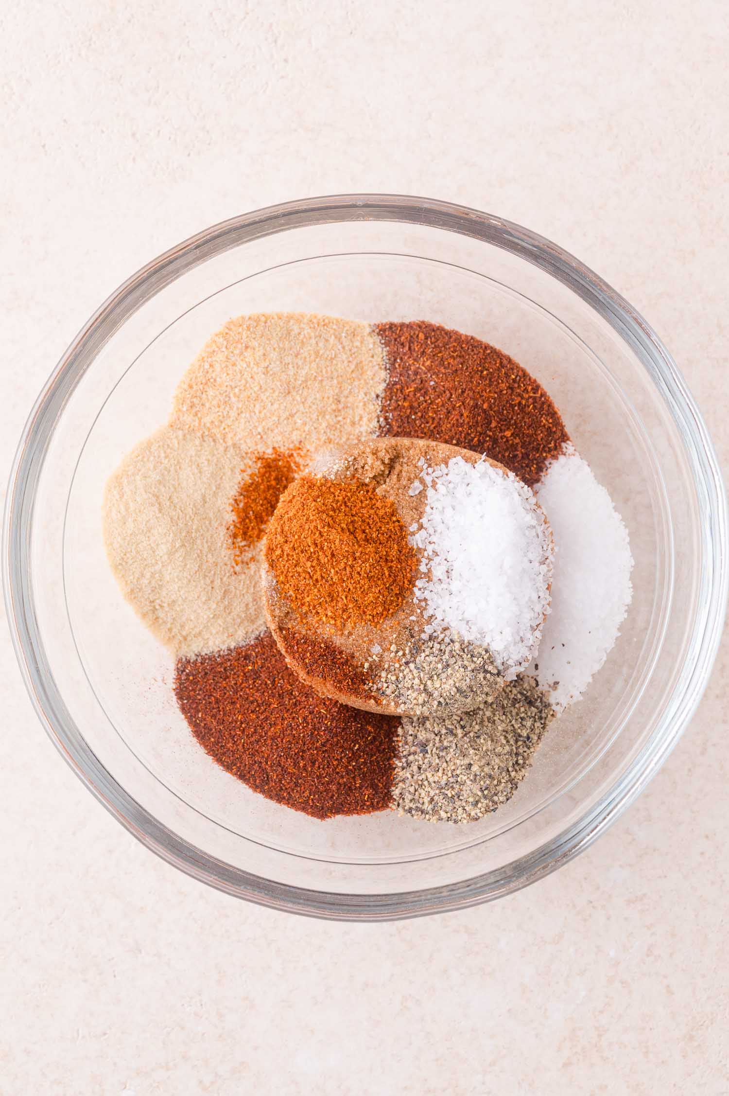A clear glass bowl containing separate piles of various spices, including salt, pepper, paprika, garlic powder, onion powder, and other seasonings, on a light surface.