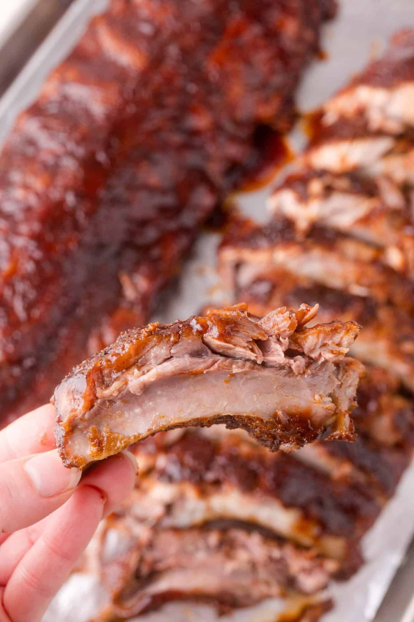 A hand holds a single, saucy barbecued rib with more sliced ribs and a whole rack in the background on a tray.