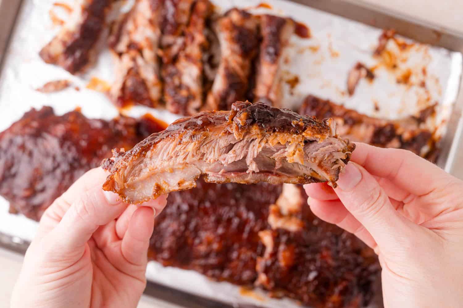 A person holds a single barbecued rib in front of a tray with more cooked ribs on parchment paper.