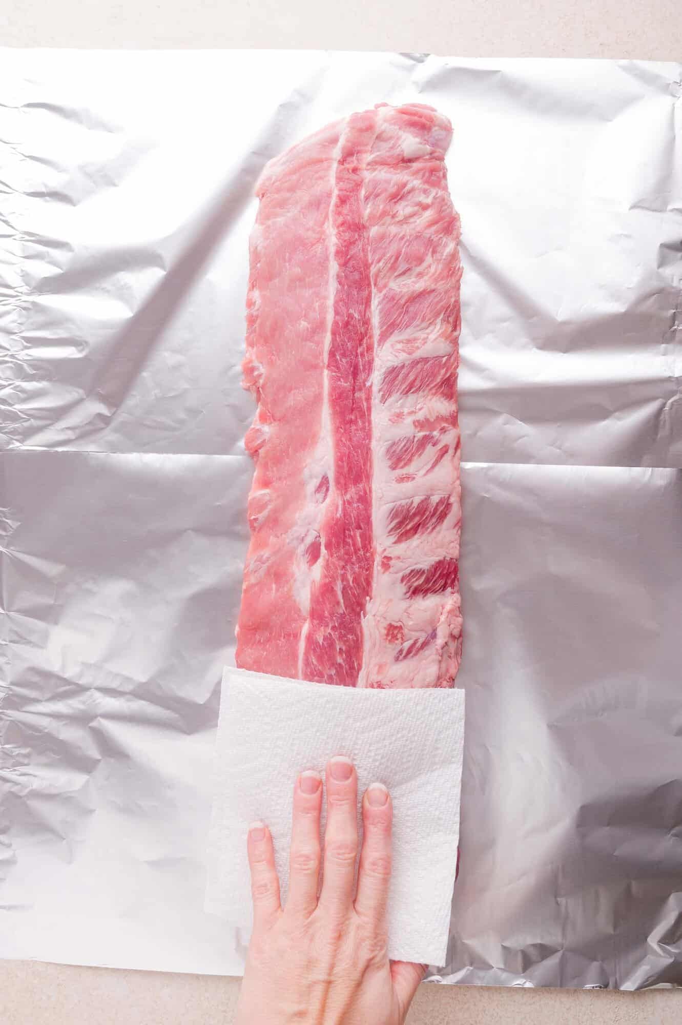 A hand using a paper towel to pat dry a raw rack of ribs placed on a sheet of aluminum foil.