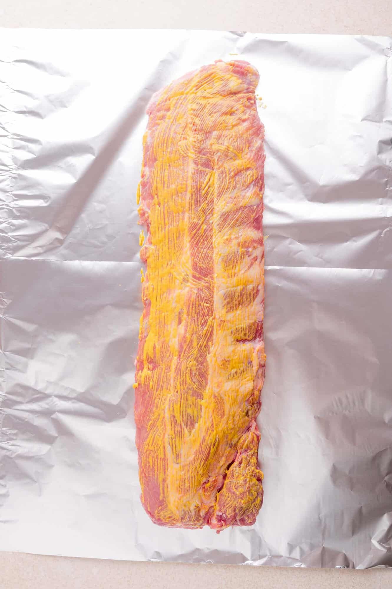 A rack of raw pork ribs coated with yellow mustard is placed on a sheet of aluminum foil.