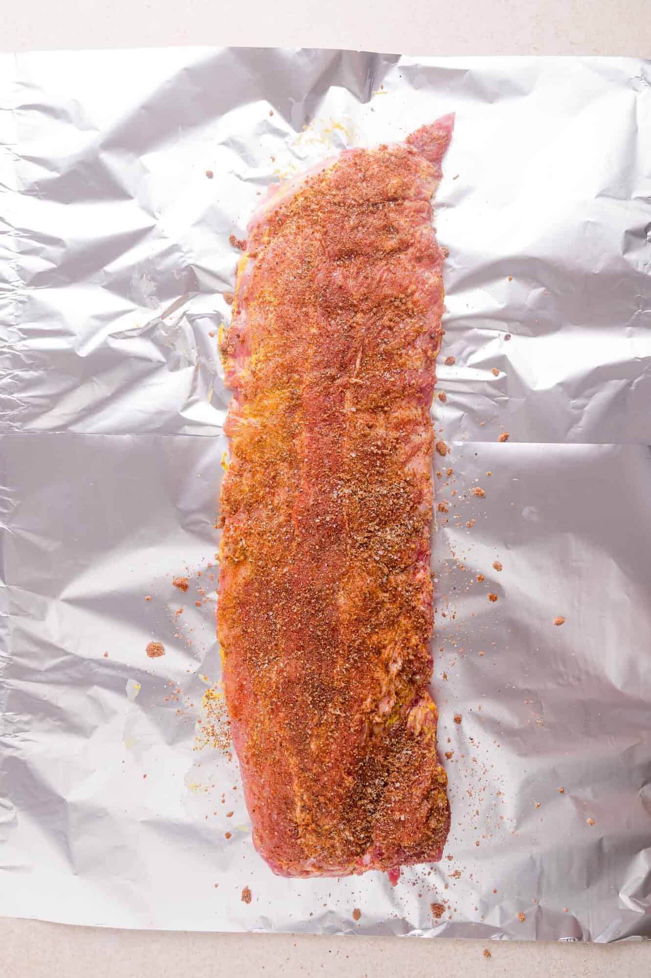 Raw pork ribs coated with dry seasoning sit on a sheet of aluminum foil, ready for cooking.